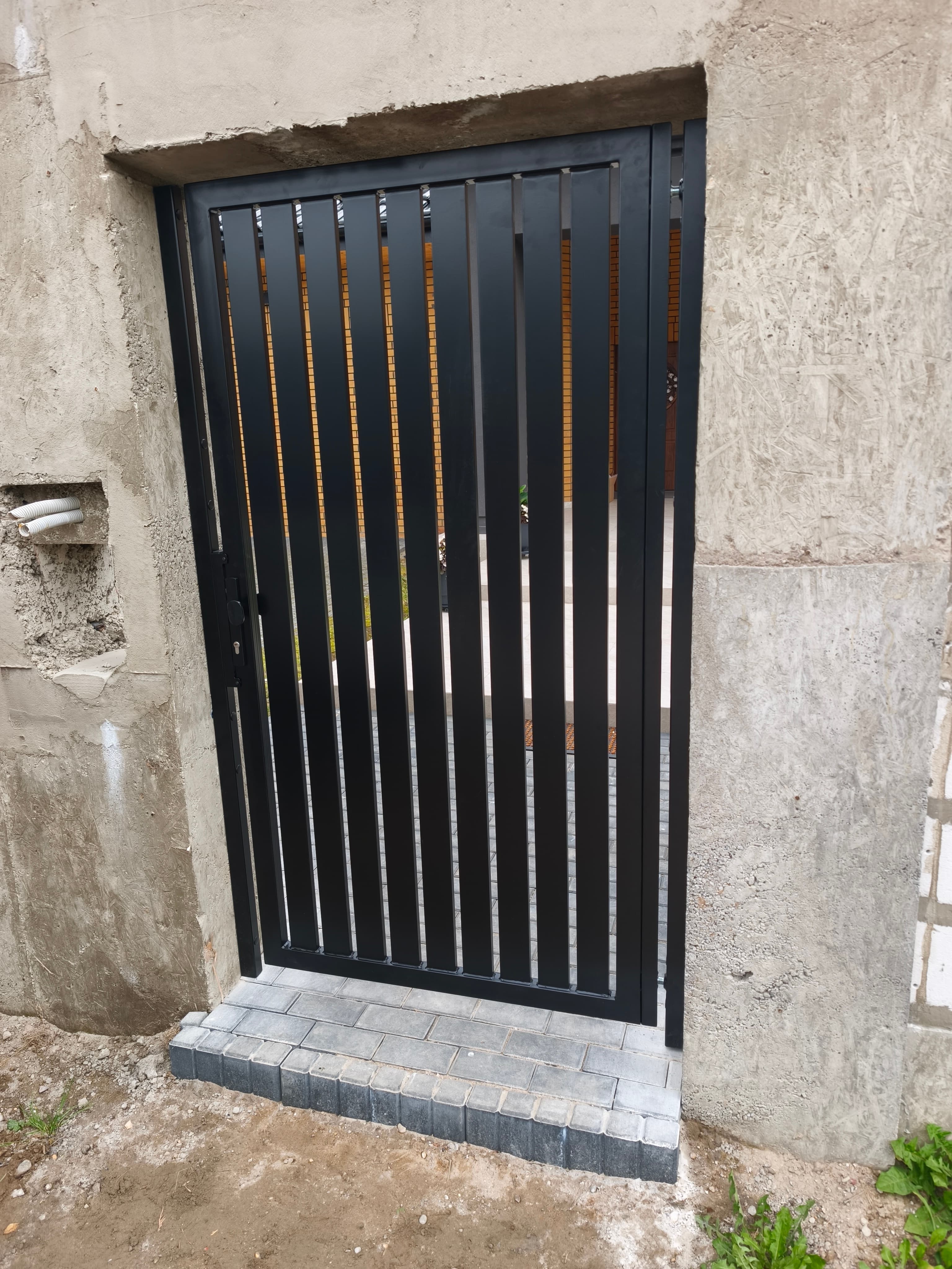Furtka wejściowa czarna z pionowymi pasami na tle betonu – montaż